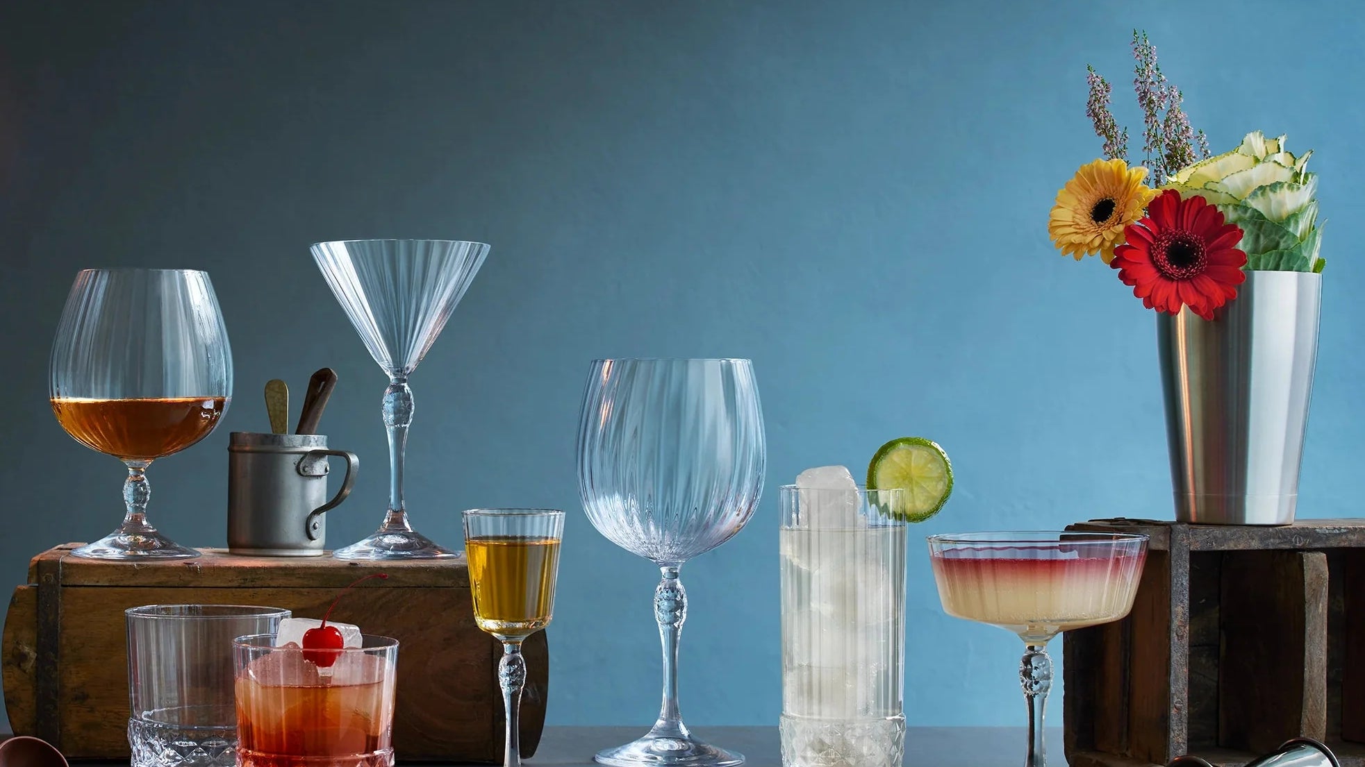 A bar-style still life with multiple clear drinking glasses and cocktail glassware arranged on a dark tabletop against a blue-gray background. Two hanging Edison bulbs illuminate the scene, with drinks, straws, a jigger.