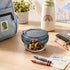 A clear glass Frigovere lunch container with a dark gray lid sits on a light wood desk, filled with mixed nuts and dried fruit. The scene includes a light blue lunch bag with a dinosaur patch, a white insulated bottle with a rainbow, crayons, a note card reading 'Have a great day!', and notebooks in a bright school-lunch setting.