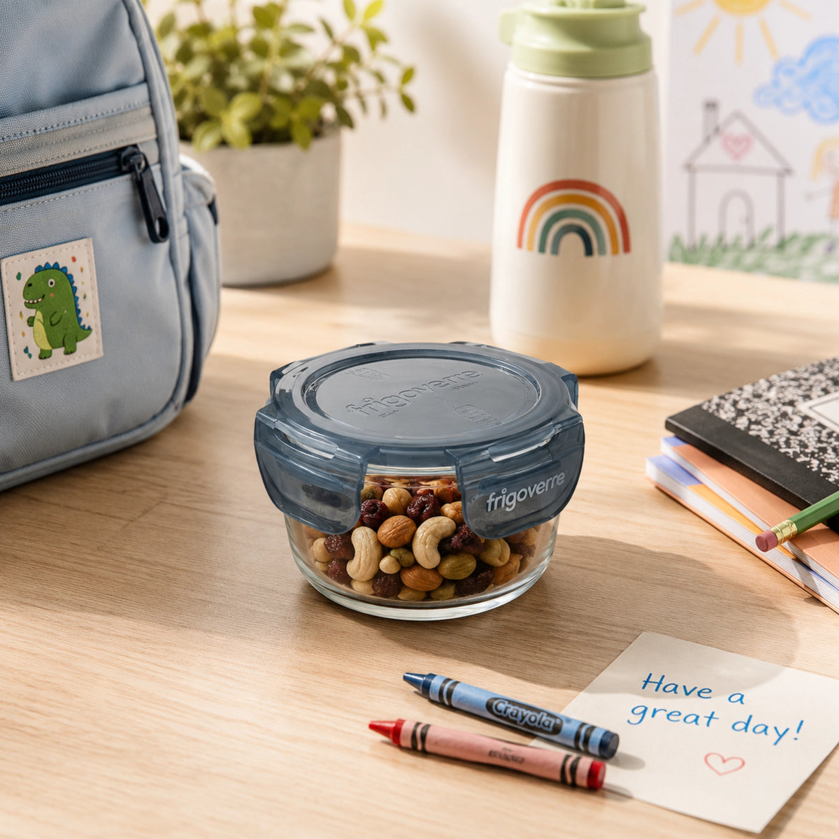 A clear glass Frigovere lunch container with a dark gray lid sits on a light wood desk, filled with mixed nuts and dried fruit. The scene includes a light blue lunch bag with a dinosaur patch, a white insulated bottle with a rainbow, crayons, a note card reading 'Have a great day!', and notebooks in a bright school-lunch setting.