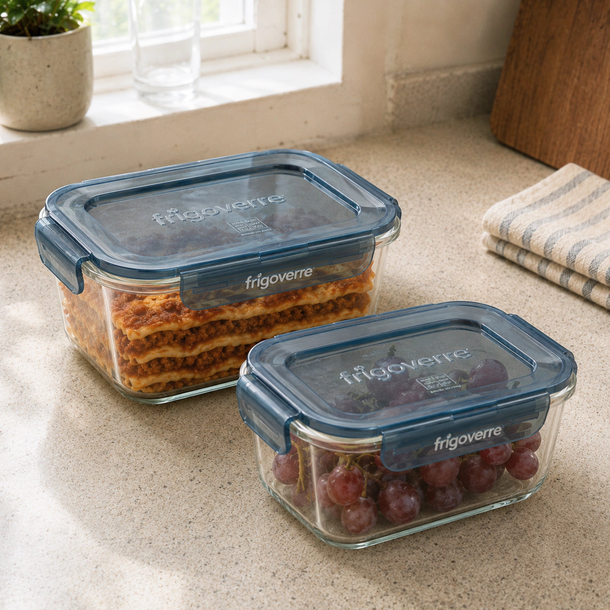 Two clear glass food storage containers with dark blue lids from frigoverre Evolution, one filled with layered lasagna and the other with red grapes, sitting on a beige kitchen countertop near a window.