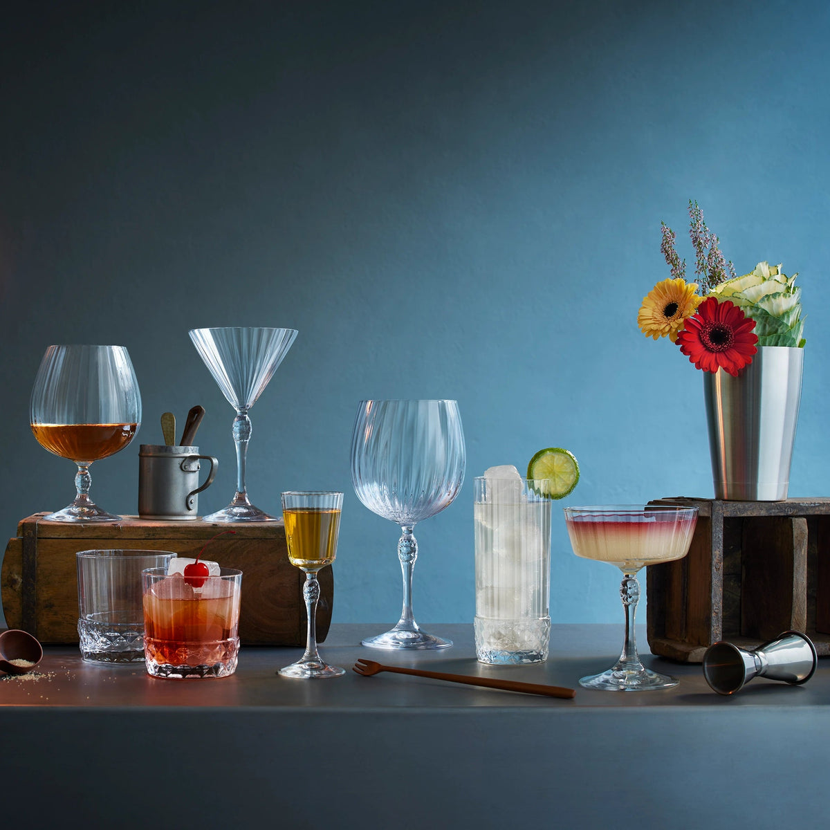 A bar-style still life with multiple clear drinking glasses and cocktail glassware arranged on a dark tabletop against a blue-gray background. Two hanging Edison bulbs illuminate the scene, with drinks, straws, a jigger, flowers in a vase, and wooden blocks used as styling props.