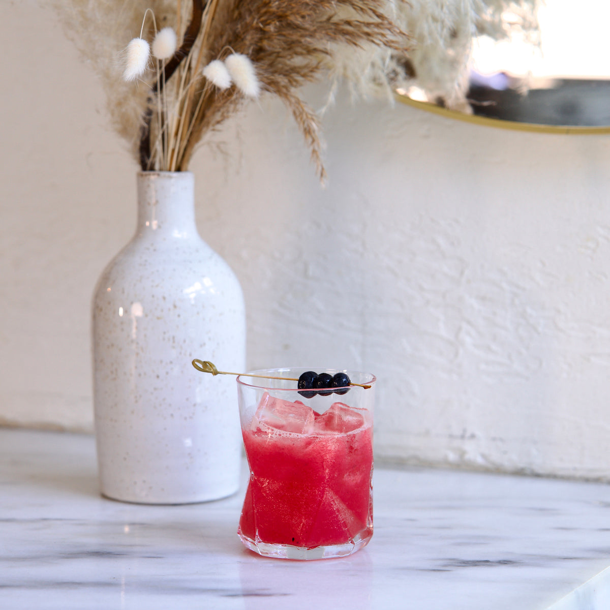 Pink cocktail with berries in a Cassiopea DOF glass, next to a white vase with dried plants on a marble surface.