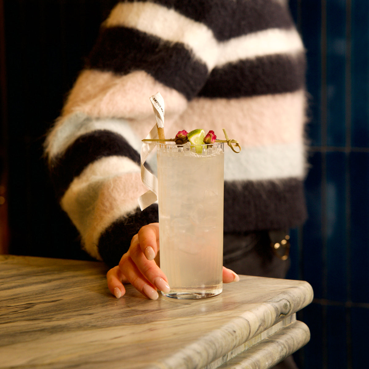 Person in striped sweater holding Exclusiva Tom Collins cocktail glass with a drink with a colorful garnish on a marble surface.